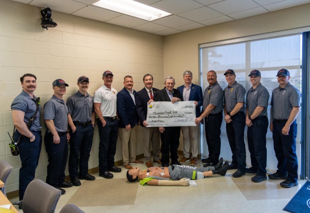 Mountain Brook Fire Department personnel and community leaders pose together holding an oversized ceremonial check for $13,600 made out to Mountain Brook Fire from Cawaco RC&D, for the purchase of an AmbuMan training manikin. An AmbuMan Advanced manikin lies on the floor in the foreground. The group is gathered inside a Mountain Brook Fire Department training room.