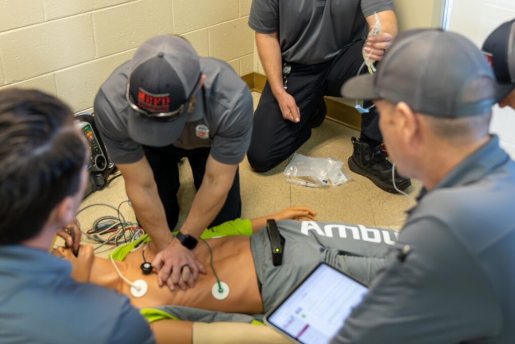 A Mountain Brook Fire Department firefighter wearing an MBFD cap performs chest compressions on an AmbuMan Advanced training manikin during a hands-on demonstration. Two other personnel kneel nearby, with one holding IV tubing and another viewing a tablet displaying real-time training data. Medical wires and training supplies are visible on the floor around the manikin.
