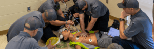 A group of Mountain Brook Fire Department personnel surrounds an AmbuMan Advanced training manikin on the floor, practicing an advanced emergency medical scenario. Personnel are performing chest compressions, managing an airway with a bag-valve mask, operating a cardiac monitor, and reviewing data on a tablet. Medical wires and equipment are connected to the manikin throughout the simulation.