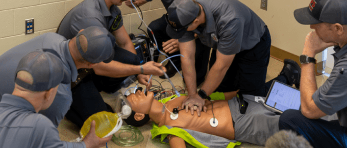 A group of Mountain Brook Fire Department personnel surrounds an AmbuMan Advanced training manikin on the floor, practicing an advanced emergency medical scenario. Personnel are performing chest compressions, managing an airway with a bag-valve mask, operating a cardiac monitor, and reviewing data on a tablet. Medical wires and equipment are connected to the manikin throughout the simulation.