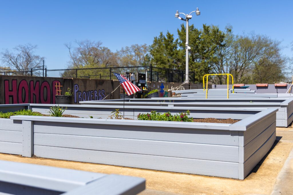 Rows of newly built gray raised garden beds stretching across the Lovelady Center rooftop under a clear blue sky, with an American flag and murals visible in the background.