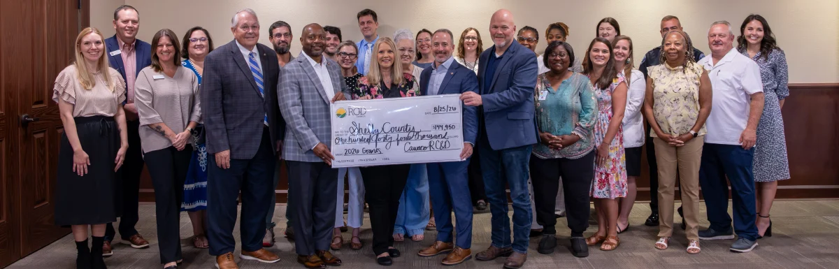Legislators and grant recipients pose in a Calera City Hall meeting room. Four legislators in the center hold an oversized check from Cawaco RC&D to Shelby County for $144,950 with the memo "2026 Grants."