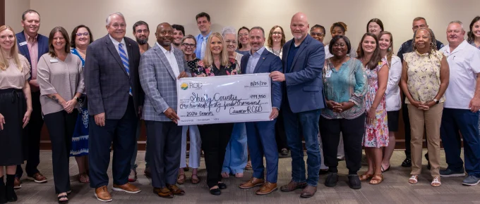Legislators and grant recipients pose in a Calera City Hall meeting room. Four legislators in the center hold an oversized check from Cawaco RC&D to Shelby County for $144,950 with the memo "2026 Grants."