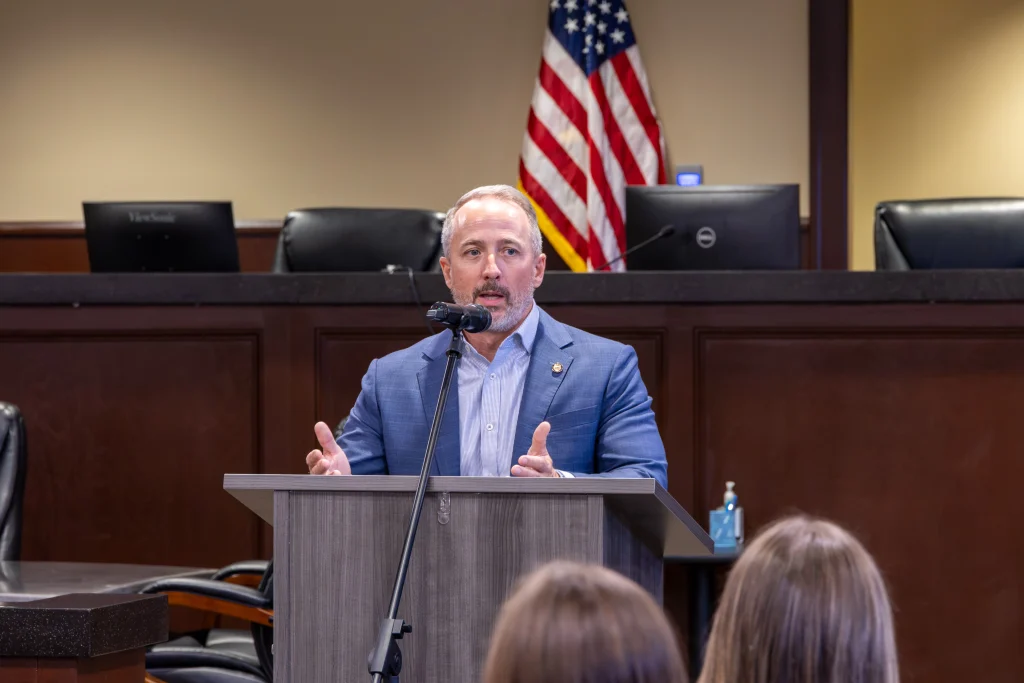Rep. Russell Bedsole speaks into a microphone at a podium, gesturing with both hands. He wears a blue jacket over a light blue shirt, with an American flag behind him.
