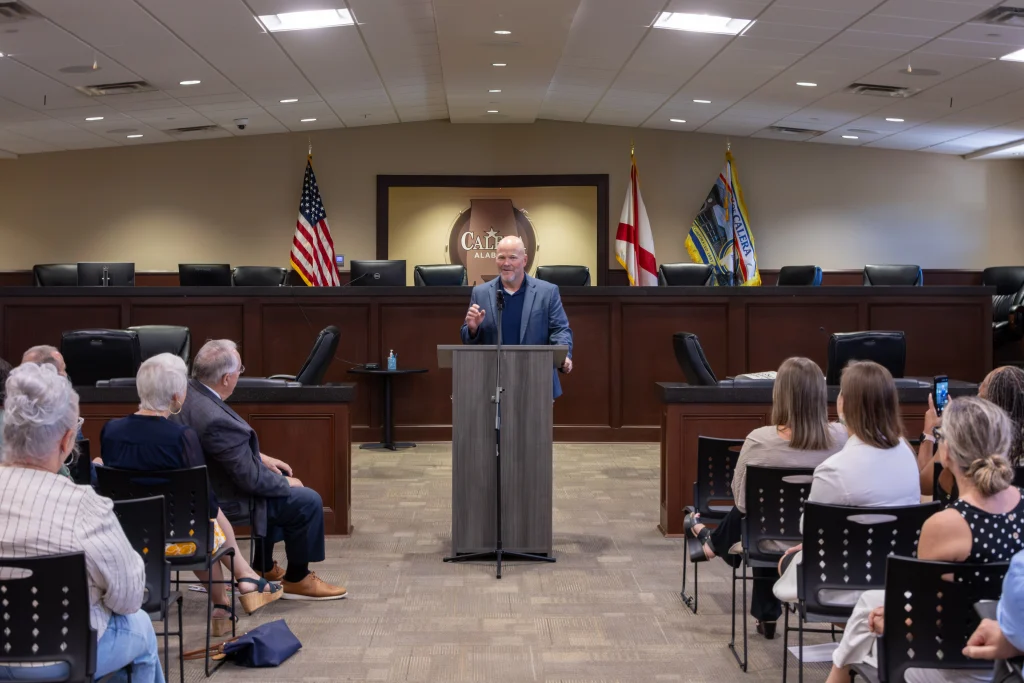 Rep. Corley Ellis speaks at a podium in the Calera council chamber. Audience members sit in the foreground, and the Calera, Alabama logo hangs on the wall behind him.