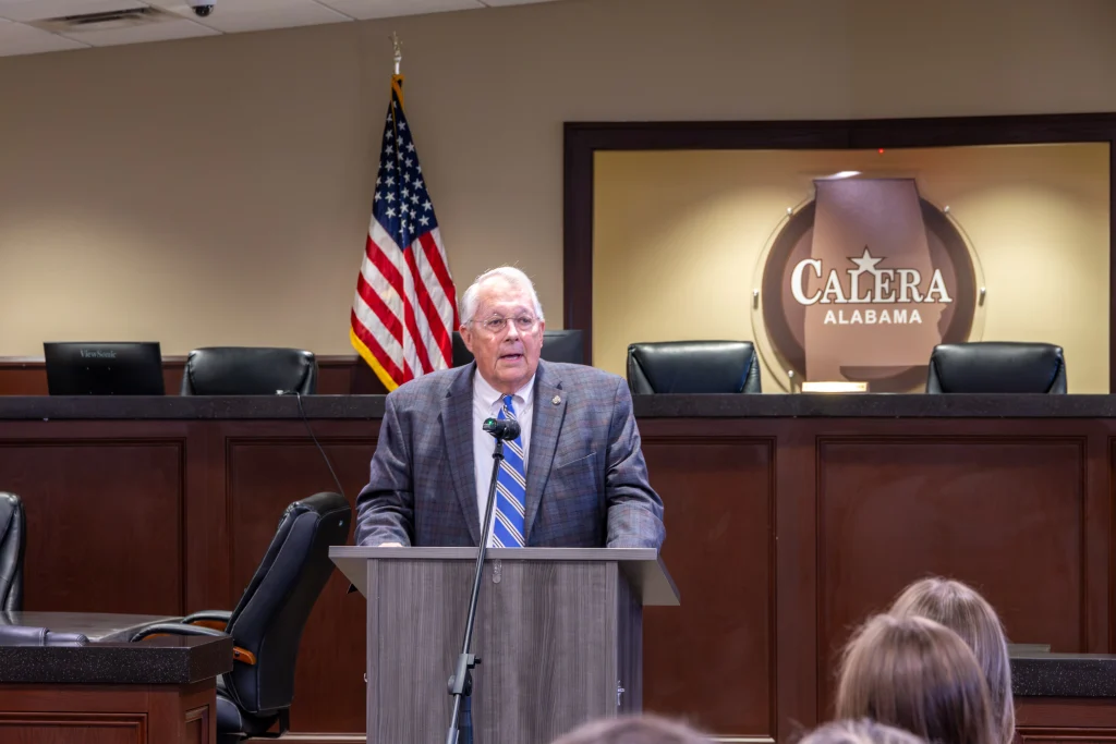 Rep. Arnold Mooney speaks at a podium in front of the Calera, Alabama logo and an American flag. He wears a gray plaid jacket and a blue striped tie.