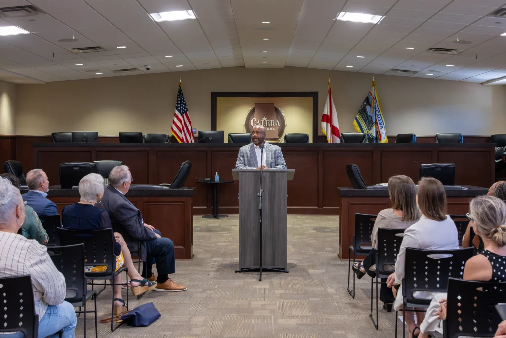 Rep. Kenneth Paschal speaks at a podium in the Calera council chamber. Audience members sit in rows of chairs in the foreground, and the American, Alabama, and Calera flags stand behind him.