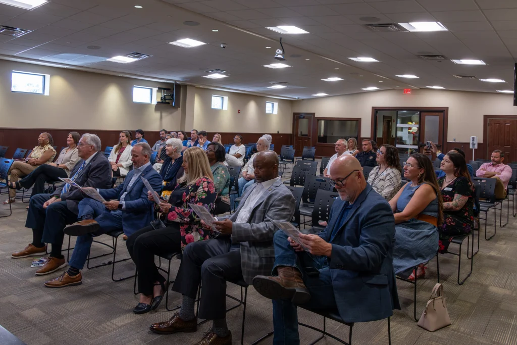 Attendees sit in rows of black chairs in the Calera City Hall meeting room, several holding printed flyers as they watch the ceremony.