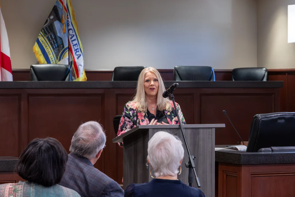 Sen. April Weaver speaks at a podium in Calera City Hall, wearing a floral blazer, with the Calera city flag behind her.
