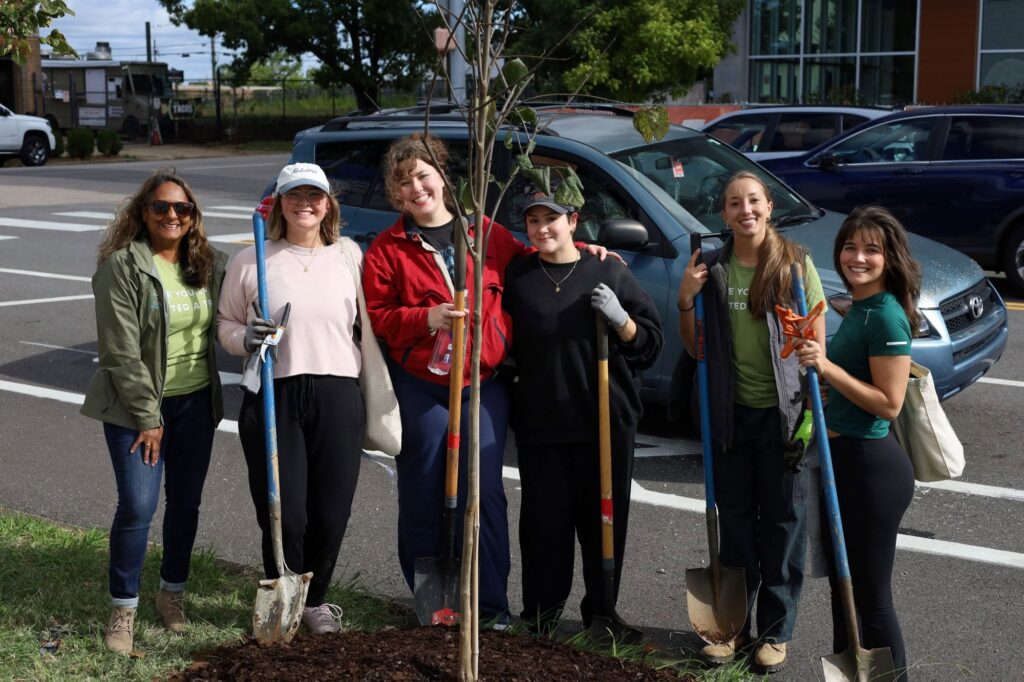 Program director Grace with five smiling volunteers stand around a newly planted sapling with fresh mulch at its base, holding shovels and pruners, with a street and parked cars behind them.