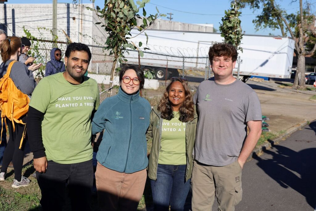 Four smiling Cool Green Trees staff pose together in front of a newly planted sapling, wearing green event shirts, a Cool Green Trees fleece, and a gray Cool Green Trees T-shirt.