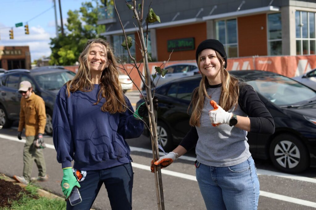 Two smiling volunteers in work gloves stand beside a newly planted sapling on a street median, one giving a thumbs-up, with parked cars and a modern building behind them.