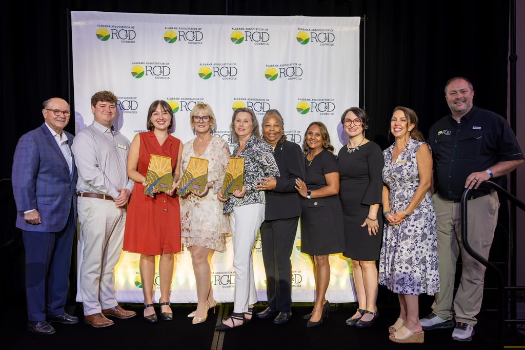 Ten people stand in a row in front of an Alabama Association of RC&D Councils backdrop, smiling at the camera. Three women in the center hold ceramic Alabama-shaped awards with a sunrise and river design.