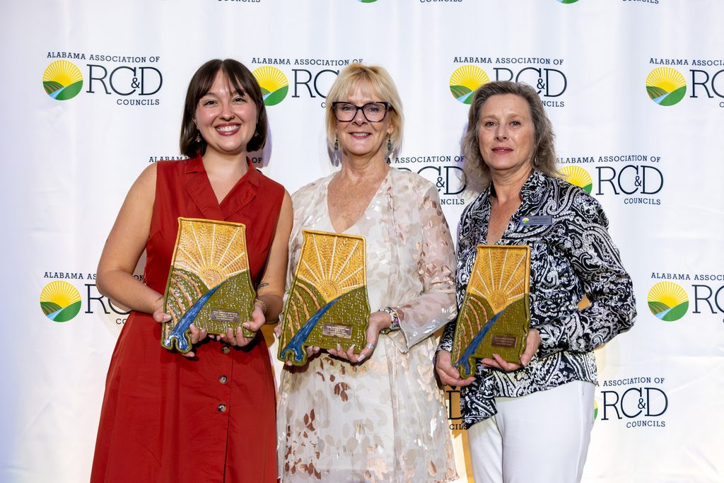 Three women from Cawaco RC&D pose in front of an Alabama Association of RC&D Councils backdrop, each holding a ceramic Alabama-shaped award decorated with a sunrise, green fields, and a blue river.