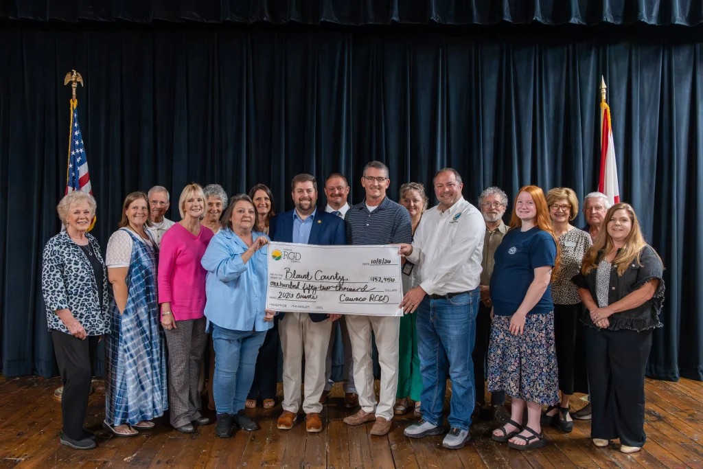 Eighteen people pose on stage at the Community Arts Council of Blount County in front of a black curtain, holding a ceremonial Cawaco RC&D check for $152,456 dated 10/8/26, with the American and Alabama flags behind them.