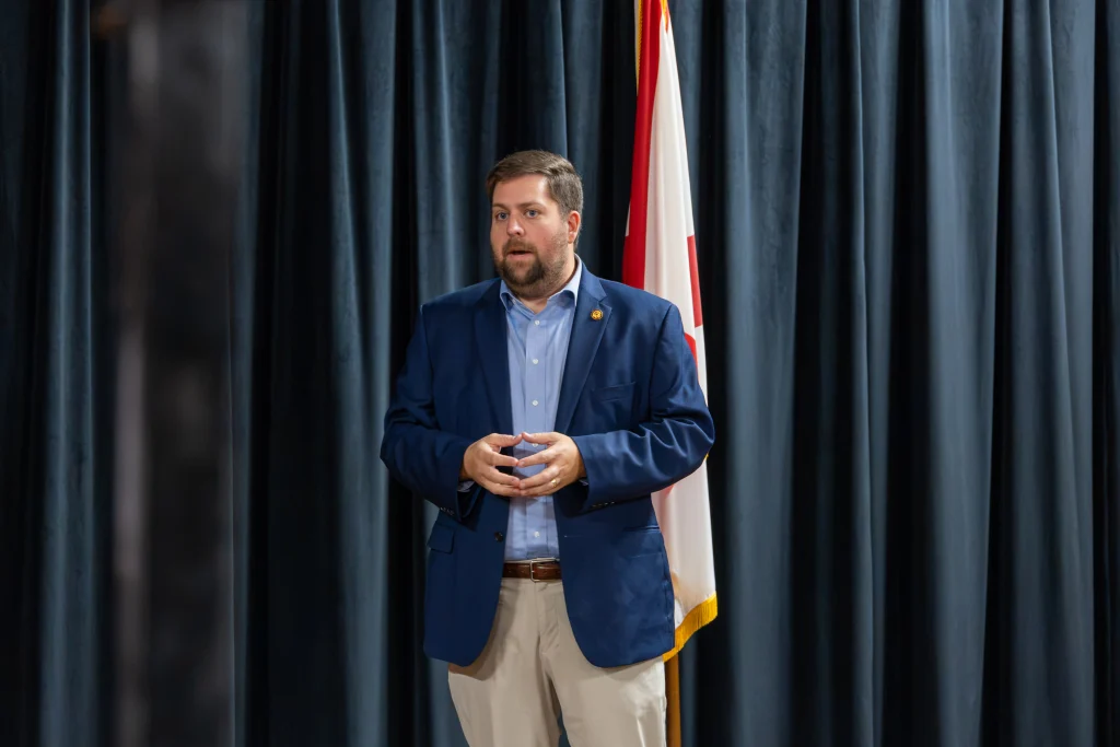 Sen Wes Kitchens, in a navy blazer and light blue shirt, speaks to the audience in front of a dark curtain and the Alabama flag.