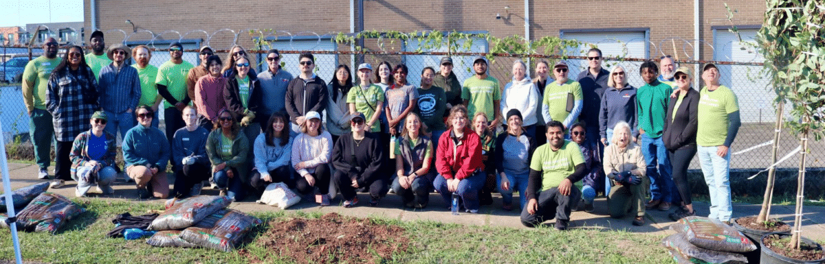 Large group of Cool Green Trees volunteers in green “Have you ever planted a tree?” shirts and casual clothes, posing together on a sidewalk beside bags of mulch, a freshly dug planting spot, and young potted trees, with a brick building and fence behind them.