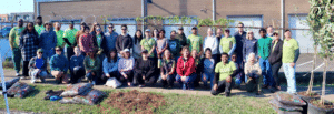 Large group of Cool Green Trees volunteers in green “Have you ever planted a tree?” shirts and casual clothes, posing together on a sidewalk beside bags of mulch, a freshly dug planting spot, and young potted trees, with a brick building and fence behind them.