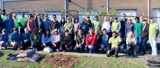Large group of Cool Green Trees volunteers in green “Have you ever planted a tree?” shirts and casual clothes, posing together on a sidewalk beside bags of mulch, a freshly dug planting spot, and young potted trees, with a brick building and fence behind them.